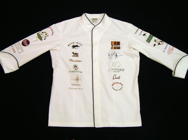 Image of Chef Coat (Chef Jacket) Style CBM103H: Shown in White Supima� gabardine, with black piping (collar, cuffs, front). Embroidered with the logos of; Le Bernardin, Picholine, Artisanal, Artisanal Cheese, Ouest, Le Cirque, Town, Country, Alto, L'Impero, A Casa, D'Artagnan, Gramercy Tavern, Food Network, Tabla, D�vi, Highlands Bar and Grill, Kerry Heffernan, Taste of T and Supima.