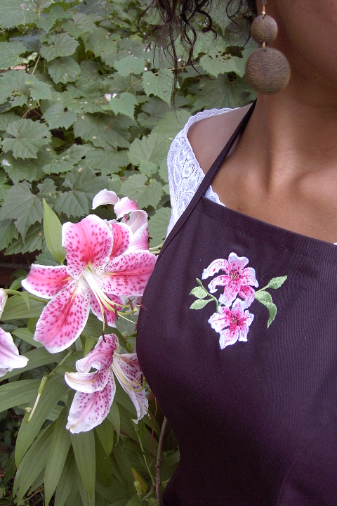 Women's Bib apron style W740; Shown in black, 100% cotton gabardine with two side hip tailored welt pockets, Ruby Glint piping on pocket welts, & embroidered stargazer lilies on center chest.