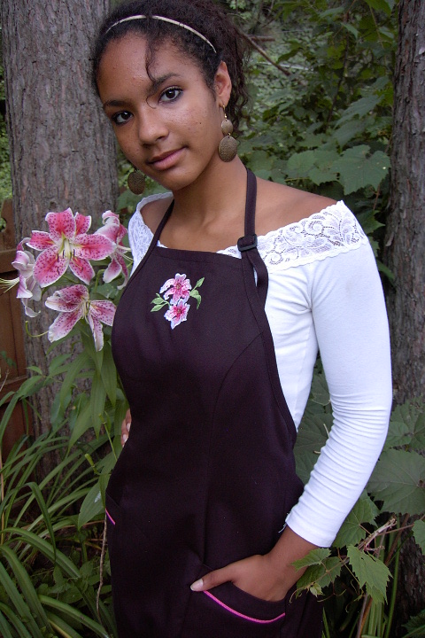 Women's Bib apron style W740; Shown in black, 100% cotton gabardine with two side hip tailored welt pockets, Ruby Glint piping on pocket welts, & embroidered stargazer lilies on center chest.