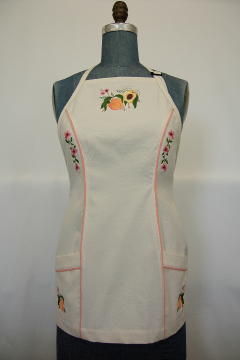 Women's Bib apron style W740 shortened 5 inches; Shown in Natural, 100% cotton riptstop with two side hip tailored welt pockets, bisque piping on pocket welts & along princess seams, embroidered peach botanical on center chest, embroidered peach blossom chains; one along each princess seam & individual peaches; one on each pocket.