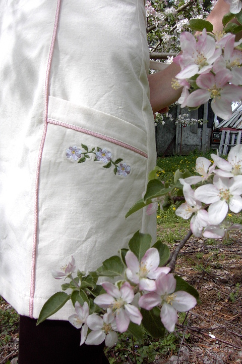 Close up image of embroidered apple blossom chain embroidered on Women's Bib apron style W740; Shown in Natural, 100% cotton riptstop with two side hip tailored welt pockets, pink sham piping on pocket welts & along princess seams, embroidered apple botanical on center chest & embroidered apple blossom chains; one on each pocket.