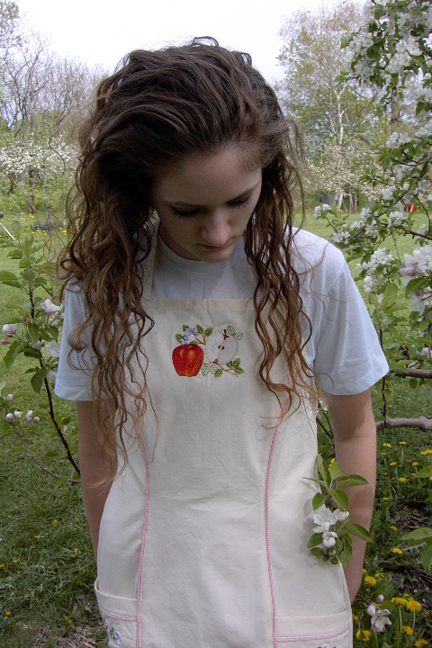 Women's Bib apron style W740; Shown in Natural, 100% cotton riptstop with two side hip tailored welt pockets, pink sham piping on pocket welts & along princess seams, embroidered apple botanical on center chest & embroidered apple blossom chains; one on each pocket.