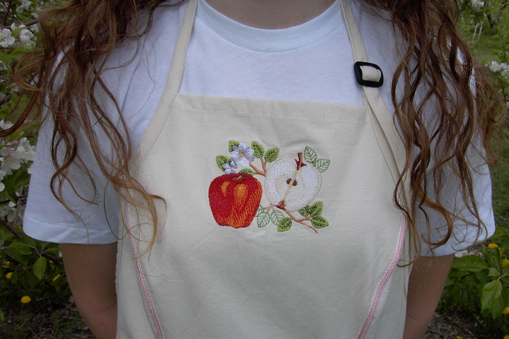 Close up image of embroidered apple botanical embroidered on Women's Bib apron style W740; Shown in Natural, 100% cotton riptstop with two side hip tailored welt pockets, pink sham piping on pocket welts & along princess seams, embroidered apple botanical on center chest & embroidered apple blossom chains; one on each pocket.