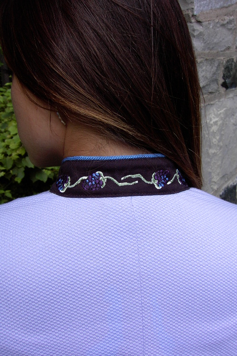 Close up image of blackberry chain embroidery along the collar of Women's Chef Coat style BSW104; Shown in Lavender & black, 100% cotton petti point pique & 100% cotton Gabardine, two front hip tailored welt pockets, Copen piping (collar, front, cuffs & pockets), Black Gabardine, hand tied knot buttons & blackberry embroideries on the chests, pockets, & back of collar.