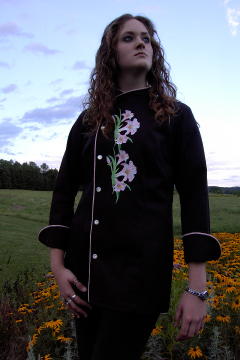 Women's Chef Coat Style CBW105: Shown in Black 100% cotton Supima® gabardine, with a left sleeve tailored welt pocket, Pink Sham piping (cuffs, collar, front), faux Mother-of-Pearl buttons & embroidered Easter Lily clusters.